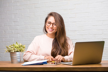 Portrait of young student latin woman sitting on her desk cheerful and with a big smile, confident, friendly and sincere, expressing positivity and success