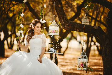 Beautiful and fashionable young bride with trendy hairstyle in stylish wedding lace dress sits on the hanging swings, decorated with flowers, and posing at the garden