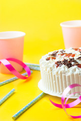 Sponge layer birthday cake with whipped cream frosting milk chocolate star sprinkles strawberry jam. Pink paper drinking cups blue straws curly ribbon on yellow background. Party celebration kids fun