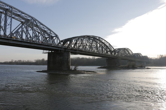 The Ernest Malinowski Bridge in Torun. Railway bridge on the Vistula river. Visible spans, steel trusses, supports, water current. A winter, cold, windy day. A shore with trees in the background.