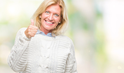Middle age blonde woman wearing winter sweater over isolated background doing happy thumbs up gesture with hand. Approving expression looking at the camera with showing success.