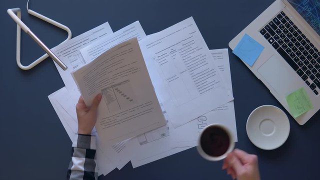 business woman in a gray shirt parses working papers sitting at the table and drinks coffee. on the table is a laptop. accelerated time. overhead shot