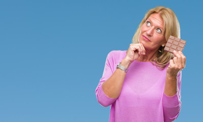 Middle age blonde woman eating chocolate bar over isolated background serious face thinking about question, very confused idea