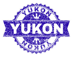 YUKON rosette stamp watermark with grunge style. Designed with round rosette, ribbon and small crowns. Blue vector rubber print of YUKON title with dirty style.