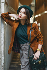 Outdoor fashion portrait of young woman wearing trendy corduroy blazer, green beret, checkered trousers, golden wrist watch, carrying leather bag,  posing in street of European city