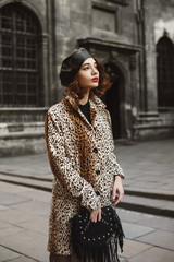 Outdoor fashion portrait of young woman wearing trendy leopard print coat, leather beret, holding...