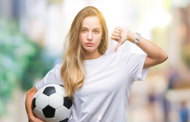Young beautiful blonde woman holding soccer ball over isolated background with angry face, negative...