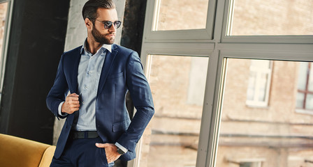 Confident and stylish. Businessman in sunglasses and fashion suit is thoughtfully looking in window while standing at office - obrazy, fototapety, plakaty