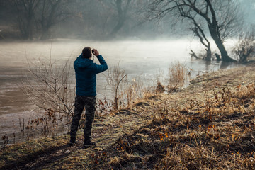 Young man watching a birds and morning fog with binoculars near river