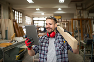 Young professional carpenter holding wood material and looking at tablet in carpentry workshop.