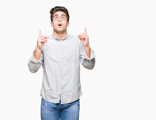 Young handsome man wearing glasses over isolated background amazed and surprised looking up and pointing with fingers and raised arms.