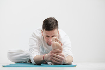 Obraz premium Handsome young man in white clothes does exercises. Yoga asanas and poses for stretching and meditating on a blue rug. Against the background of a white wall.
