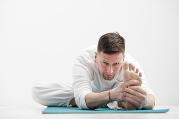 Fototapeta premium Handsome young man in white clothes does exercises. Yoga asanas and poses for stretching and meditating on a blue rug. Against the background of a white wall.