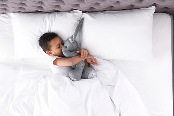 Cute little African-American boy with toy rabbit sleeping in bed, top view. Space for text