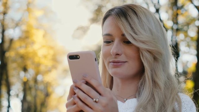 Close up portrait of cute girl wearing white sweater using smart phone and browsing on mobile phone in city park. Portrait of woman writing message on her smartphone at sunset.