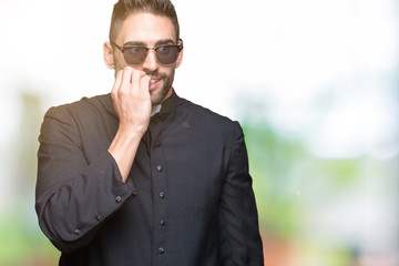 Young Christian priest wearing sunglasses over isolated background looking stressed and nervous with hands on mouth biting nails. Anxiety problem.