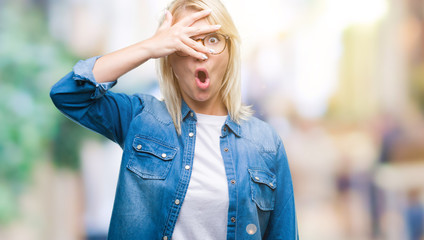 Young beautiful blonde woman wearing glasses over isolated background peeking in shock covering face and eyes with hand, looking through fingers with embarrassed expression.