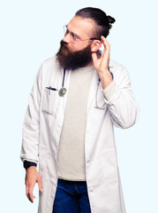 Young blond doctor man with beard wearing medical coat smiling with hand over ear listening an hearing to rumor or gossip. Deafness concept.