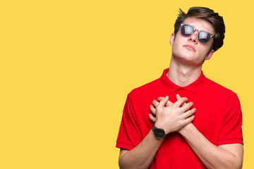 Young handsome man wearing sunglasses over isolated background smiling with hands on chest with closed eyes and grateful gesture on face. Health concept.