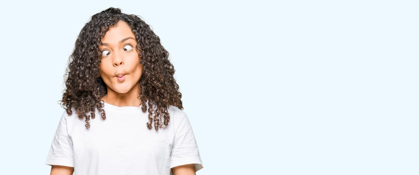 Young Beautiful Woman With Curly Hair Wearing White T-shirt Making Fish Face With Lips, Crazy And Comical Gesture. Funny Expression.