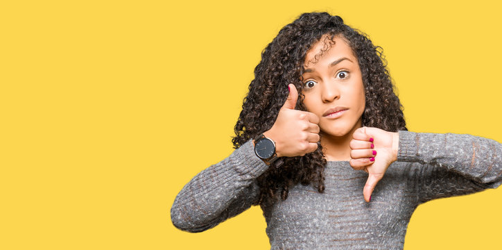 Young beautiful woman with curly hair wearing grey sweater Doing thumbs up and down, disagreement and agreement expression. Crazy conflict