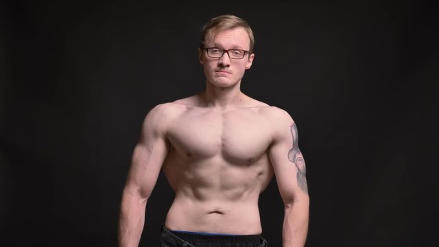 Portrait of naked young muscular man in glasses with tattoo watching calmly into camera on black background