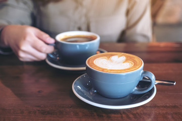 Closeup image of a hand holding a blue cup of hot black coffee with another latte coffee cup on wooden table in cafe