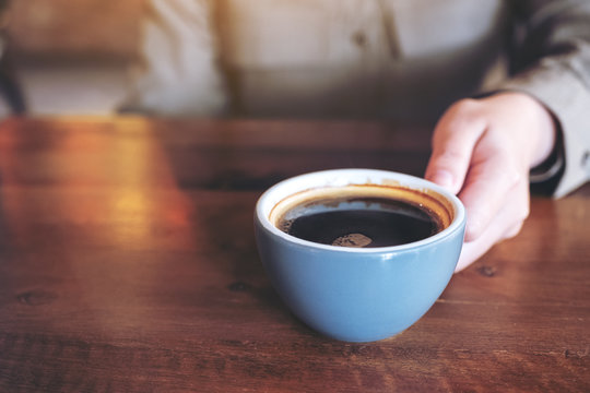 Closeup image of a hand holding a blue cup of hot black coffee on wooden table in cafe