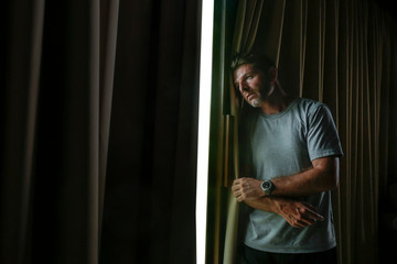 dramatic light indoors portrait of young sad and depressed attractive man looking through home room window thoughtful and pensive in pain suffering depression problem