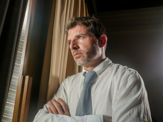office portrait of businessman working late looking worried through window thoughtful and pensive feeling sad and depressed thinking and considering business problem