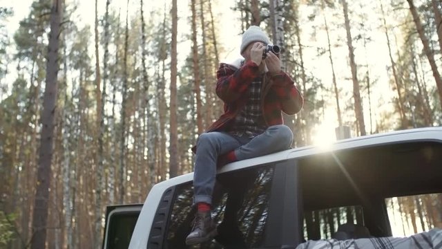 Young man in beanie sitting on car roof and taking photos of nature with camera while enjoying time in forest in autumn