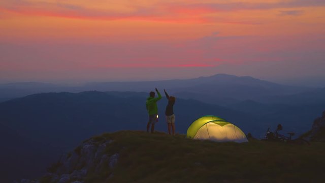 SLOW MOTION: Young man holding a lantern high fives his happy girlfriend on a cold summer evening as they enjoy a romantic camping date in the serene mountains. Carefree couple enjoy the sunset.