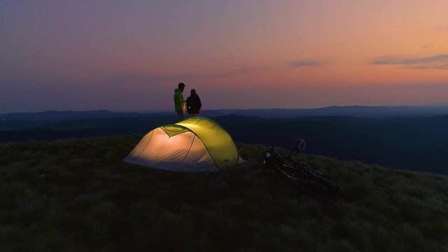 AERIAL: Young man and woman enjoying a date in the mountains on a calm summer morning. Flying around sporty couple on active vacation standing by their tent and talking on a picturesque evening.
