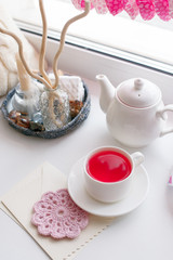 Still life interior details, cup of scented hibiscus tes, teapot, envelope, paper craft galand, candle on white windowsill. Romance morning concept