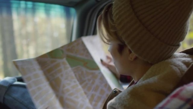 Shaky shot of young Caucasian woman using map while traveling by car and talking to unrecognizable driver
