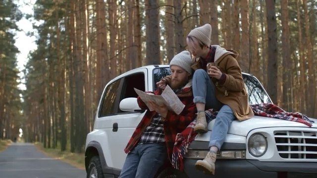 Young couple relaxing on forest road during car trip, woman drinking hot chocolate or tea and man studying map and talking to girlfriend