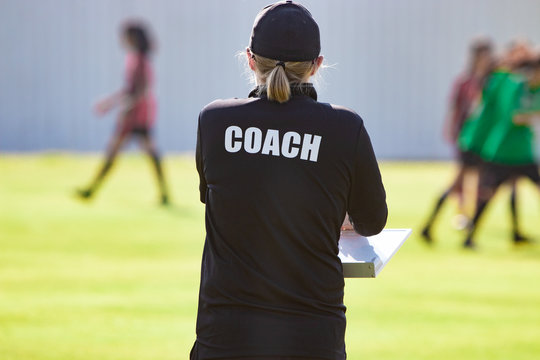Back view of female football coach in black COACH shirt at an outdoor sport field watching her team play