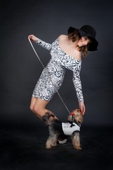 young beautiful woman in black hat with yorkshire terrier on leash in photo studio stands on black background