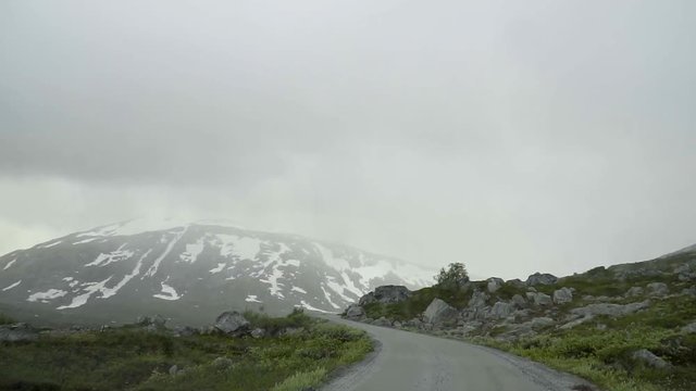 Driving through the Old Strynefjell Mountain Road in Norway with bad weather conditions and mountains with snow in the background in slow motion.