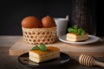 layer vanilla flavoured cake place on black plate on the wooden table there are white plate and coffee bean in glass bottles and bucket of water ceramic and basket of eggs place backside.