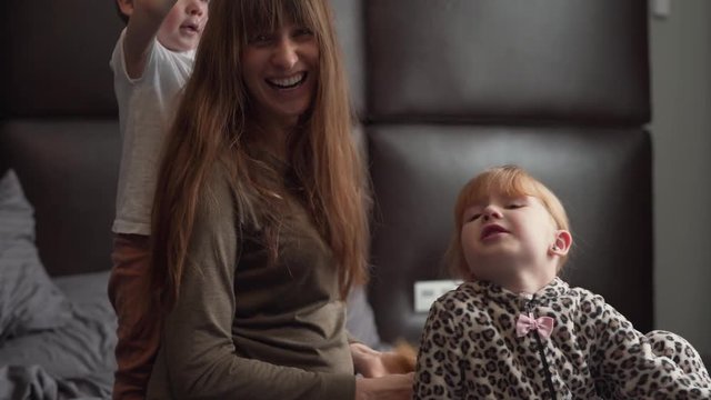 Young pregnant woman is making a braid from her daughter's hair. They are sitting on a bed. Her son is perfumed his mom and everyone is laughing.