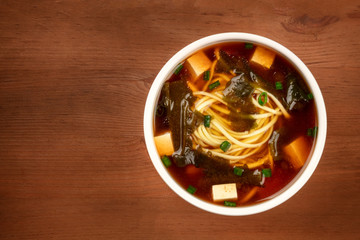 A bowl of miso shiru soup with tofu, scallions, noodles, and wakame seaweed, shot from the top on a rustic wooden background with copy space