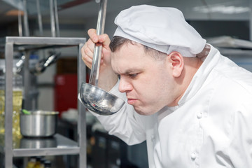 Obraz na płótnie Canvas Closeup portrait man professional chef in white uniform is trying dish made out of kitchen ladle. Concept of quality control in the restaurant, the development of new menu, an intern chef