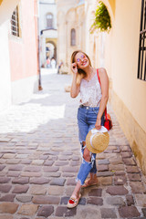 Beautiful woman in blue jeans walks through the streets of the old town with a straw hat and pink sunglasses. Attractive blonde girl with beautiful smile.