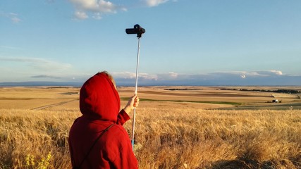 Young boy recording a video of agrarian landscape. Fields sown with wheat and barley growing as far as the eye can see on the horizon. Natural blue sky totally calm in a summer afternoon.