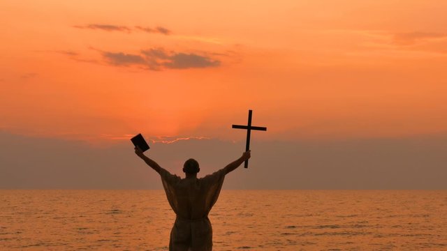 4К. Man ascetic, monk take cross  and Bible up  against sunset on seacost. Christian  team