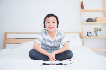 Young asian boy wearing headset listening music and play games from tablet computer on bed