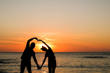 Front view of a full body of couple silhouettes making a love symbol with their hands on  a date at sunset on the beach