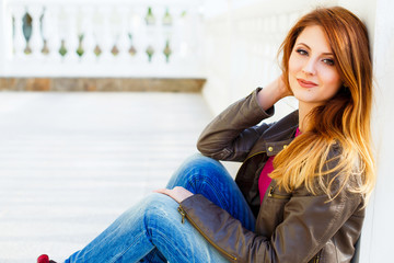 Beautiful young stylish woman with red hair, wearing jeans and leather jacket sitting on the stairs. Casual outfit. Lifestyle portrait.