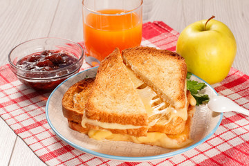 Toasted slices of bread with cheese and green parsley on white plate, glass of orange juice, apple and bowl with strawberry jam.